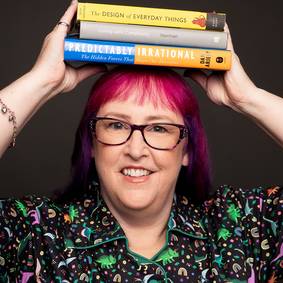 Jenni Merrifield holds three books balanced on her head,'The Design of Everyday Things', 'Living with Complexity', and 'Predictably Irrational'. She is smiling at the camera, wearing purple-rimmed glasses and a top decorated with colorful dinosaurs, rainbows, and stars.